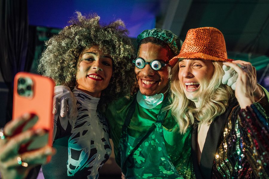 Tre vuxna poserar för en selfie på en Halloween-fest, en kvinna i skelettkostym, en man utklädd till läskig kirurg med roliga glasögon och en kvinna med paljettbeströdd hatt, vilket illustrerar önskefotos Halloween-citat för grupper.