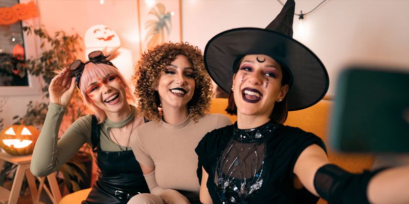Tre kvinnor i maskeradkläder på en Halloween-fest, en häxa, ett djur och en cyberpunk, poserar för en selfie i telefonen, vilket introducerar önskefotos 105 Halloween-citat för Instagram.