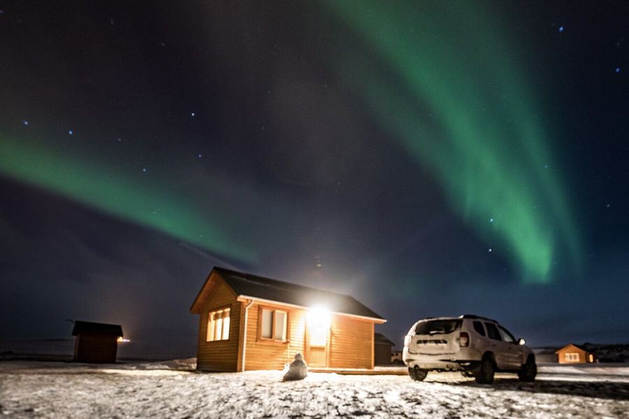 En stuga med ljus som lyser ut, bredvid en bil i ett mycket snöigt landskap, med gröna norrsken på himmelen ovanför, som illustrerar önskefotos förklaring av vad norrsken är och vad som orsakar aurora borealis.
