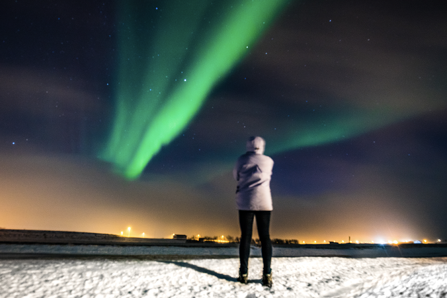 En person som står i ett snöigt landskap och tittar upp på de gröna norrsken på himlen, som illustrerar önskefotos tips för att fota norrsken med mobil.