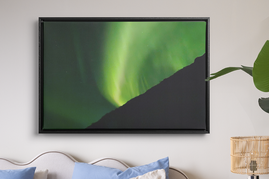 En inramad tavla med mycket grönt norrsken hänger på väggen ovanför en säng och en rafialampa, som illustrerar önskefotos väggkonst för att förvandla dina norrskensfoton till något värt att behålla.