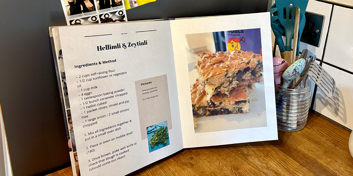 En öppen personlig kokbok ligger i ett kök, med en burk med skedar och köksredskap i bakgrunden, vilket introducerar önskefotos guide till att skapa din egen kokbok.