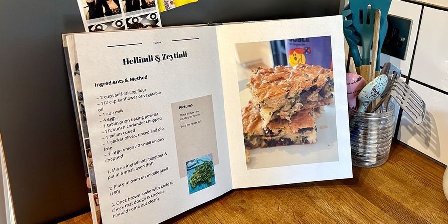 En öppen personlig kokbok ligger i ett kök, med en burk med skedar och köksredskap i bakgrunden, vilket introducerar önskefotos guide till att skapa din egen kokbok.