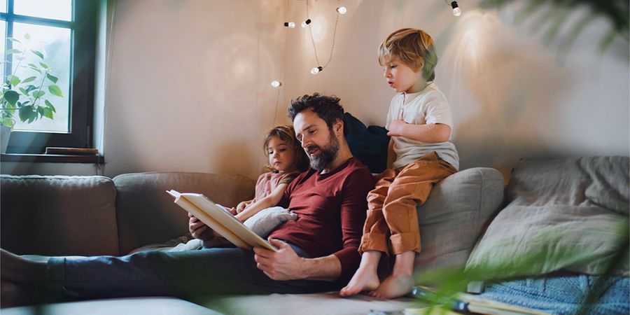 En pappa läser en bok för en liten pojke och en liten flicka i soffan, med varma ljusslingor framför dem, vilket introducerar önskefotos tips för mysig höstinredning i hygge-stil.