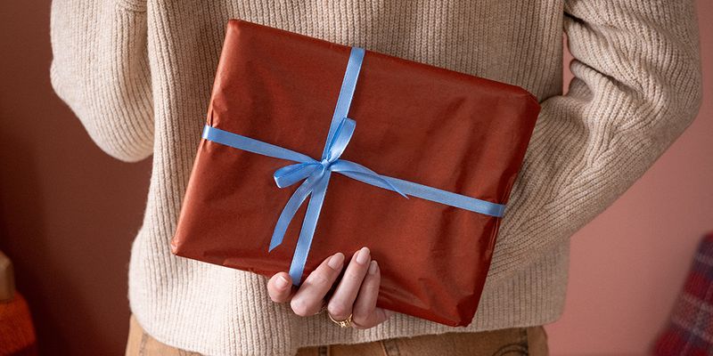 Kvinna som håller en present inslagen i brunt papper med ett blått band bakom ryggen, som illustrerar önskefotos checklista med julklappstips för en personlig fotopresent.