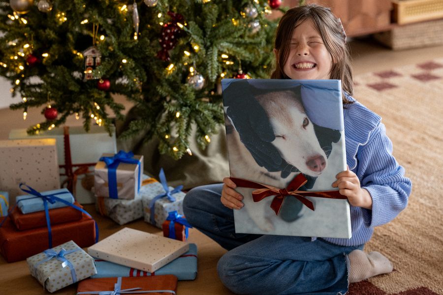 En flicka i blå tröja skrattar högt medan hon håller upp en canvastavla med en hund, med ett rött band runt, framför en julgran, vilket illustrerar önskefotos tips om väggkonst som Black Friday-julklapp.