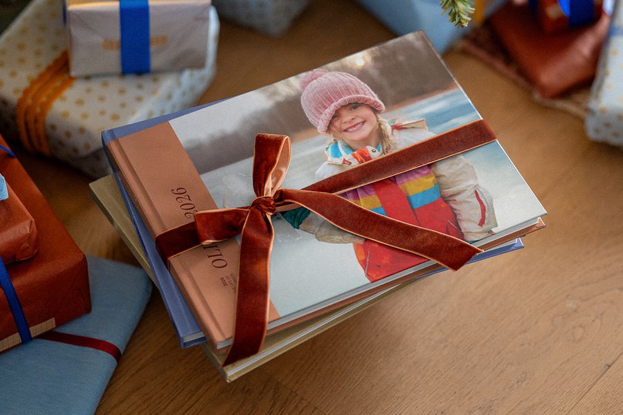 Hög med personliga fotoböcker med ett brunt sammetsband runt, med andra inslagna presenter i bakgrunden, som illustrerar önskefotos tips för att planera julklappar med personliga bilder.