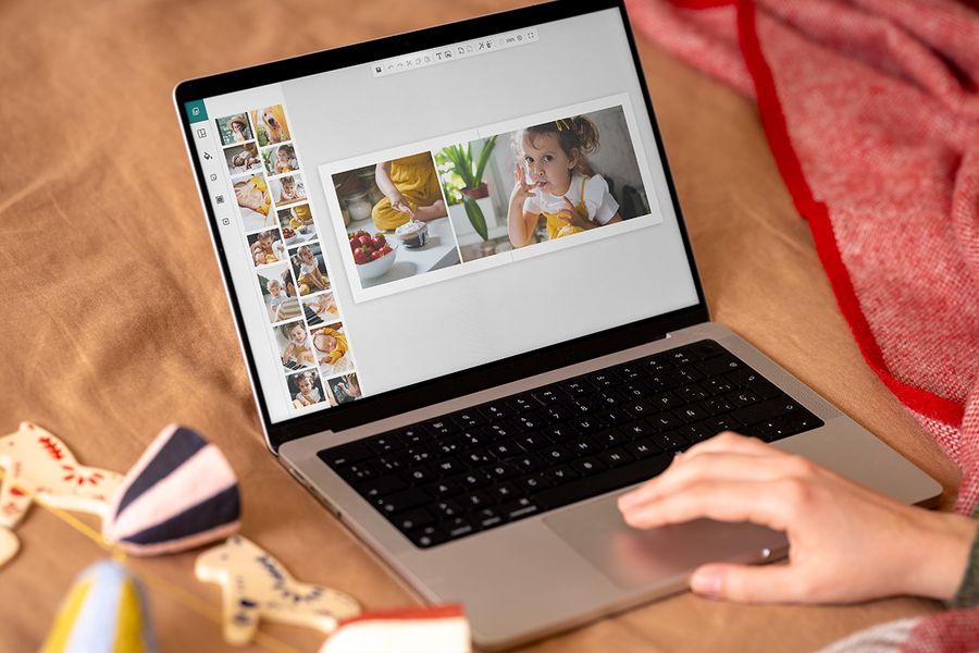 En hand på pekplattan till en bärbar dator designar en fotobok på skärmen med ett uppslag som visar en liten flicka och en skål med jordgubbar, vilket illustrerar önskefotos tips för att skapa julklappar online inför Black Friday.