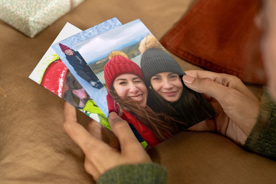 Person som håller fotoutskrifter i händerna, där den främsta visar två leende kvinnor i vintermössor och kappor, som illustrerar önskefotos guide för att hitta rätt bilder till en personlig fotopresent.