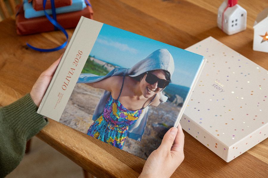 Två händer håller i en fotobok med 2026 på omslaget, där en leende kvinna i solglasögon syns på en klippig kust, bredvid en presentationsbox med konfettiprickar, vilket illustrerar önskefotos personliga fotoböcker.