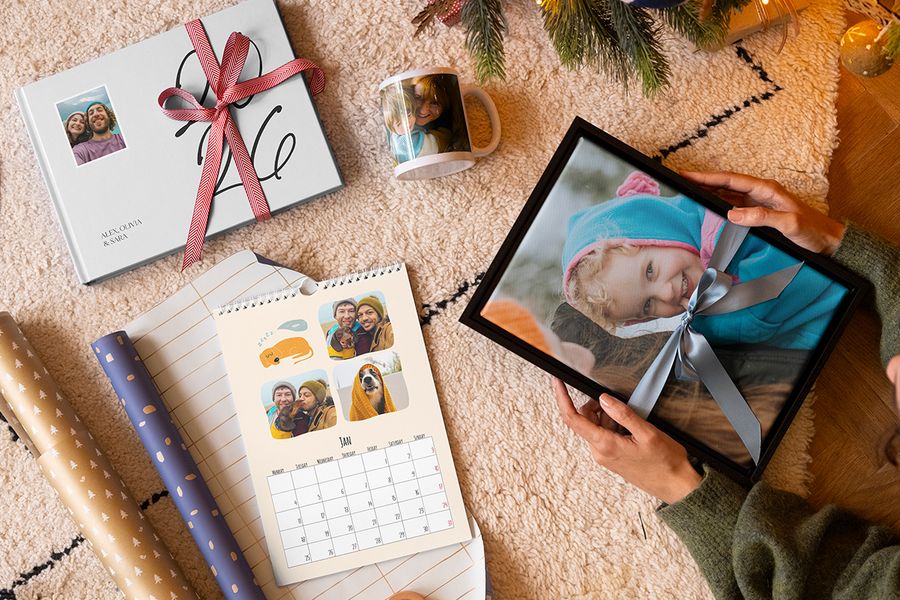 Urval av personliga presenter från önskefoto, med väggkonst av en ung flicka med ett blått band runt, en mugg med en mamma och ett barn, en personlig fotokalender och en 2026-fotobok med ett rött band runt, bredvid presentpapper och med julgransgrenar i bakgrunden, som illustrerar önskefotos tips för att fånga rätt minnen i en personlig julklapp.