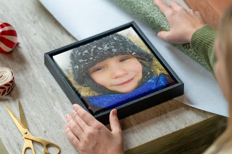 Inramad canvastavla med en ung flicka i vinterkläder i snön, med en kvinnas händer redo att slå in den, bredvid presentpapper, sax och band, som illustrerar önskefotos tips för att välja en personlig fotopresent som passar mottagarens stil.