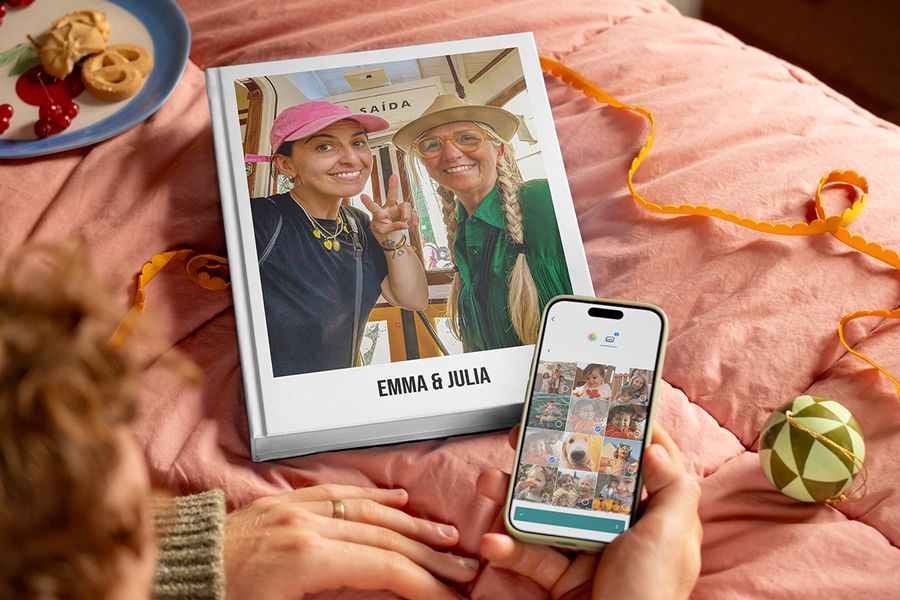 Personlig fotobok med titeln “Emma and Julia” med en mamma och hennes vuxna dotter på omslaget, bredvid en mans händer som håller en telefon för att visa hur enkelt det är att välja bilder och skapa en fotobok från önskefoto, som illustrerar önskefotos tips för att samla in bilder från familj och vänner till en personlig present.