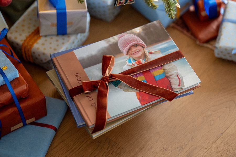 En stapel fotoböcker bundna med ett sammetsband, där den översta boken visar en leende flicka i rosa mössa och 2026 på ryggen, bland inslagna julklappar, vilket illustrerar önskefotos fotobok för året.