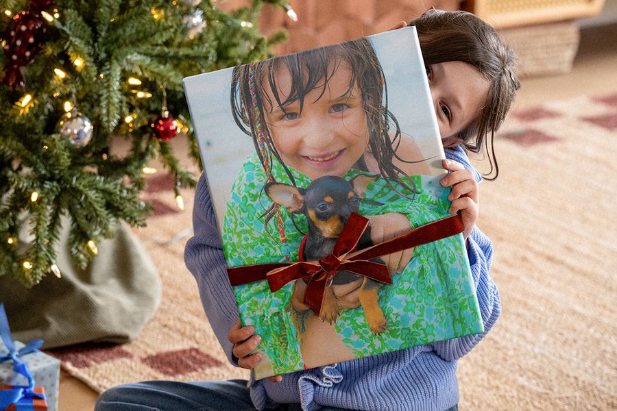 Liten flicka under julgranen som håller en stor fotobok med ett sammetsband runt och ett foto av sig själv med en chihuahua på omslaget, som illustrerar önskefotos checklista med julklappstips för en personlig fotopresent.