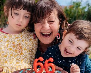 En mormor eller farmor firar sin 66-årsdag med sina två barnbarn bredvid sig och ska precis blåsa ut ljusen på tårtan, som inspiration till födelsedagspresenter från önskefotos ultimata guide till födelsedagspresenter.